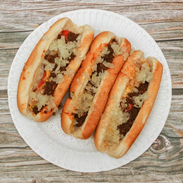 Three chili cheese wieners topped with diced onions on a white plate on a wooden table.