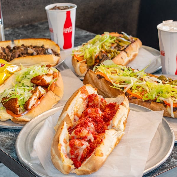 Several Cosmic Pizza Steak & Wieners subs, including meatball, cheesesteak, and chicken, with Coca-Cola cups.