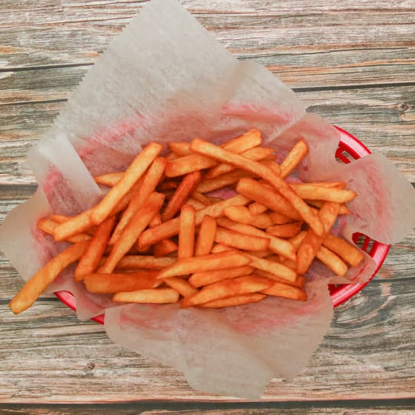 Golden-brown french fries in a red basket lined with white paper, on a rustic wooden table.